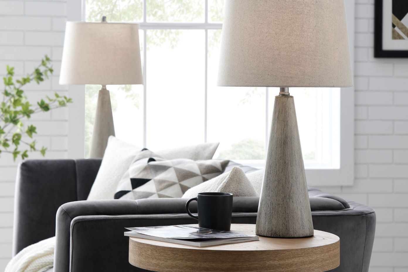 Two lamps on a side table with a black mug and a magazine. A gray sofa and pillows are in the background.
