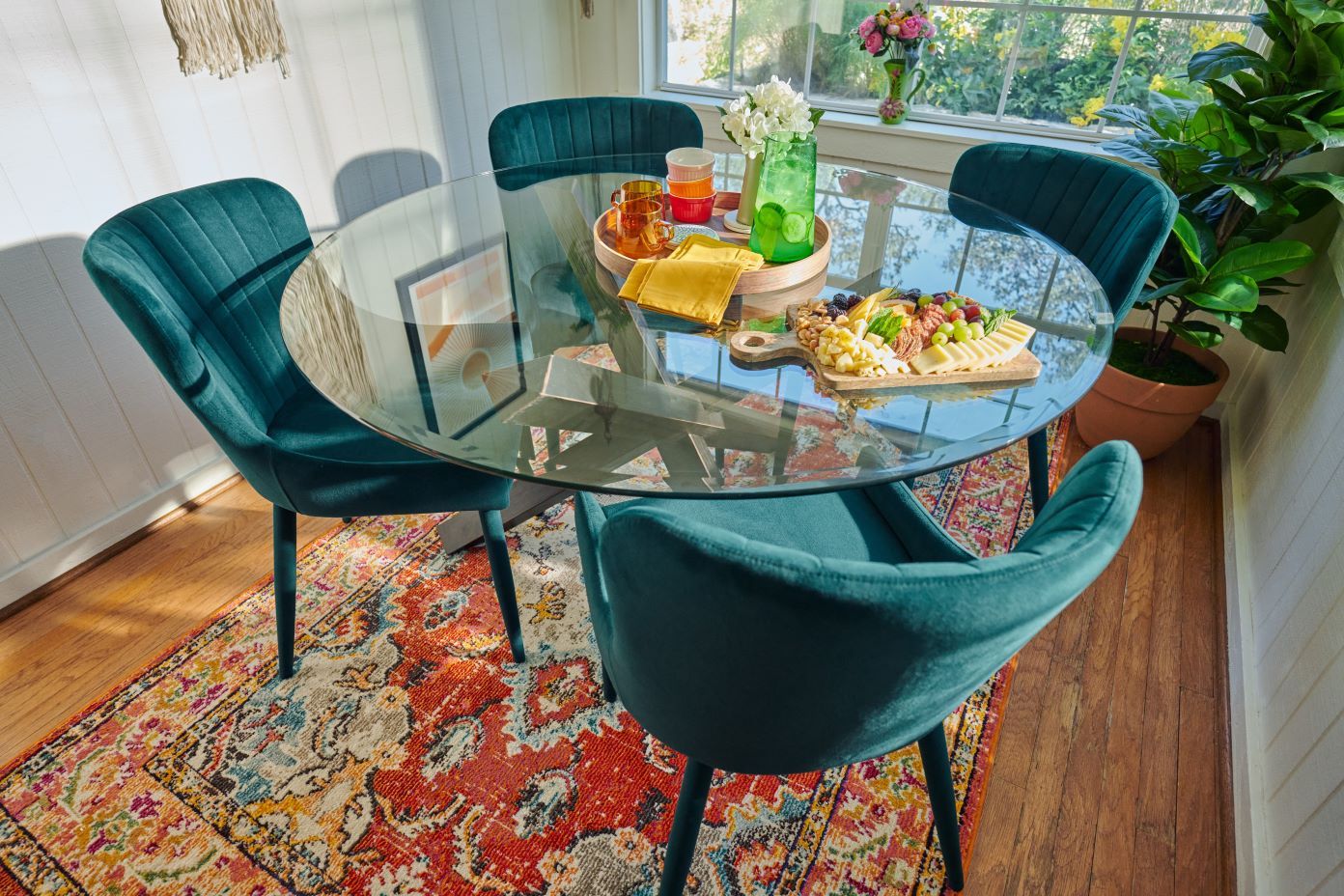 A round glass dining table with four teal velvet chairs, a colorful rug, and a charcuterie board.
