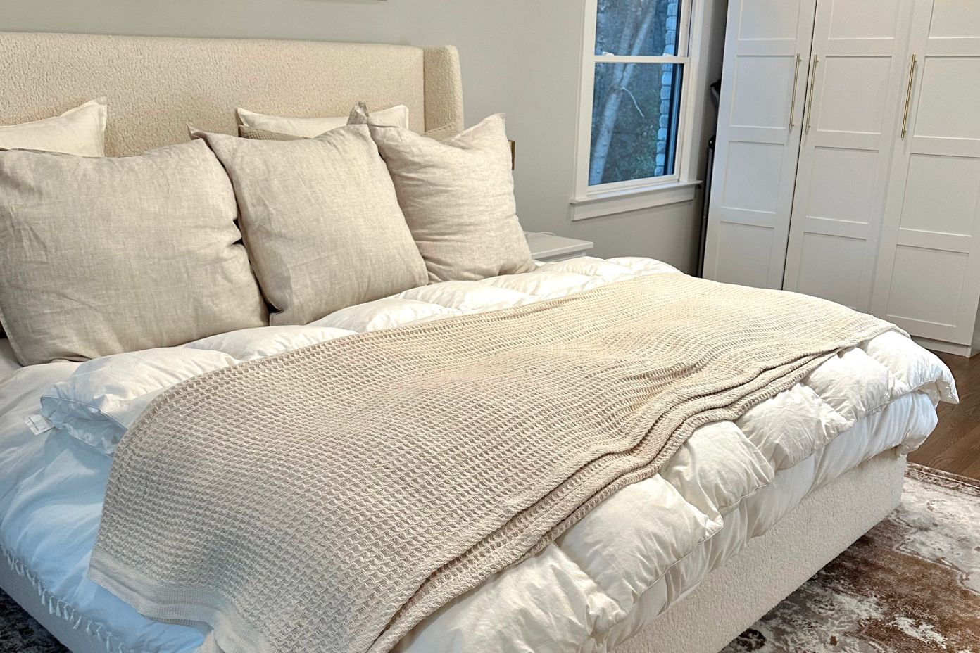 A bed with white bedding, beige pillows, and a beige waffle-knit blanket.
