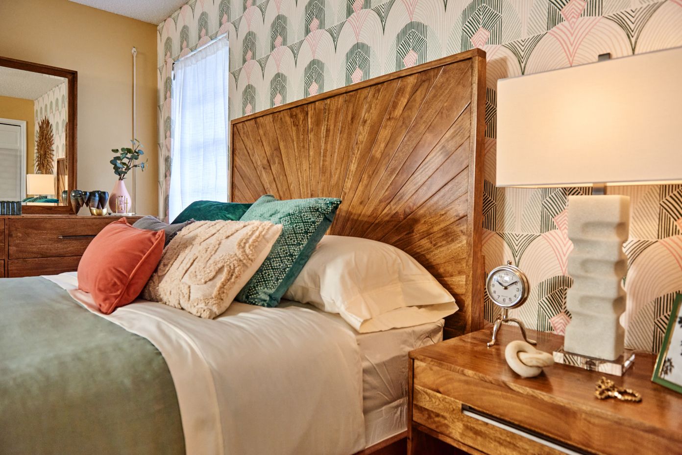 A bedroom with a wooden headboard, bed, and nightstand. There is a lamp, clock, and pillows on the bed.
