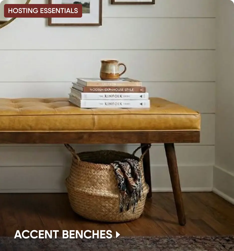 HOSTING ESSENTIALS. ACCENT BENCHES. A tufted tan leather bench with a wooden base, topped with books and a mug.
