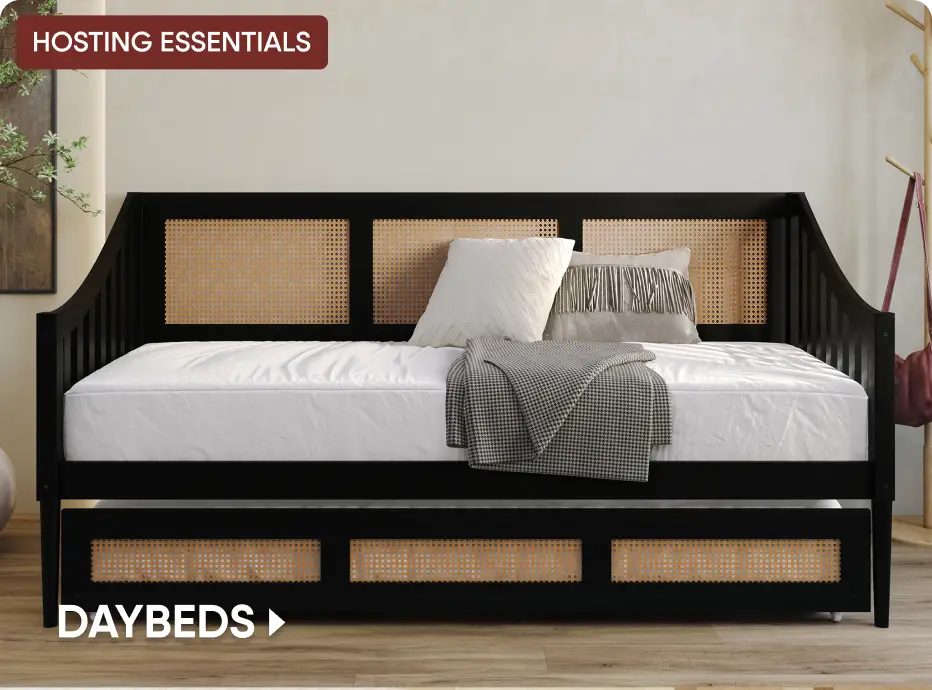 HOSTING ESSENTIALS DAYBEDS: A black wooden daybed with cane paneling and a trundle, styled with pillows and a throw blanket.