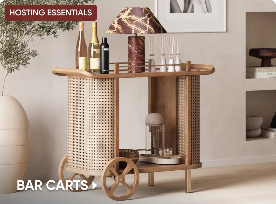 HOSTING ESSENTIALS BAR CARTS. A wooden bar cart with cane detailing, holding bottles, glassware, and a marble lamp.