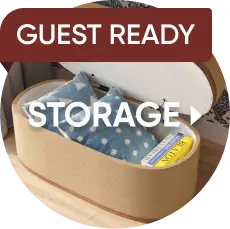 GUEST READY STORAGE: An oval, tan-colored storage ottoman with its lid open, revealing pillows and books inside.