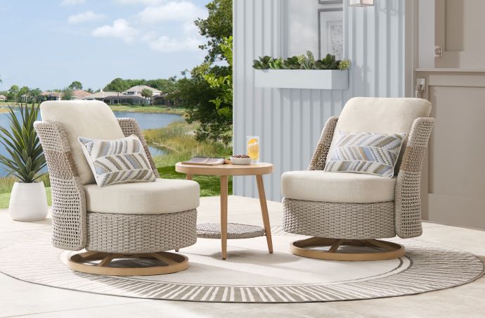 Two beige swivel chairs with pillows on a round rug, small table, and a lake view.
