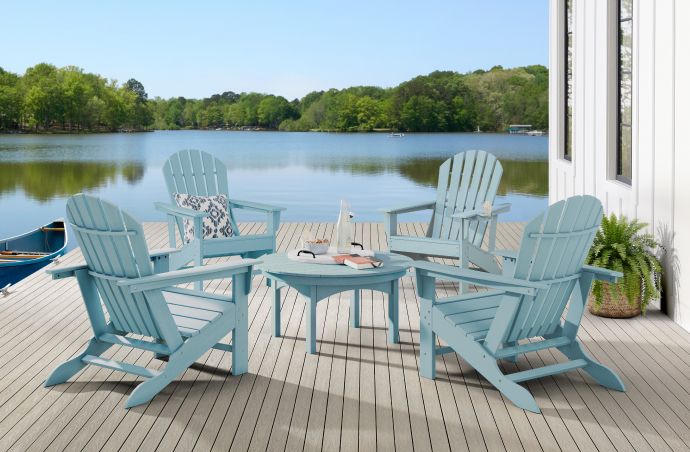Four blue Adirondack chairs and a matching table on a wooden deck overlooking a lake.
