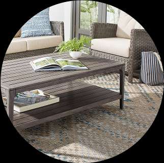 Outdoor coffee table with a lower shelf, surrounded by patio furniture and a rug.
