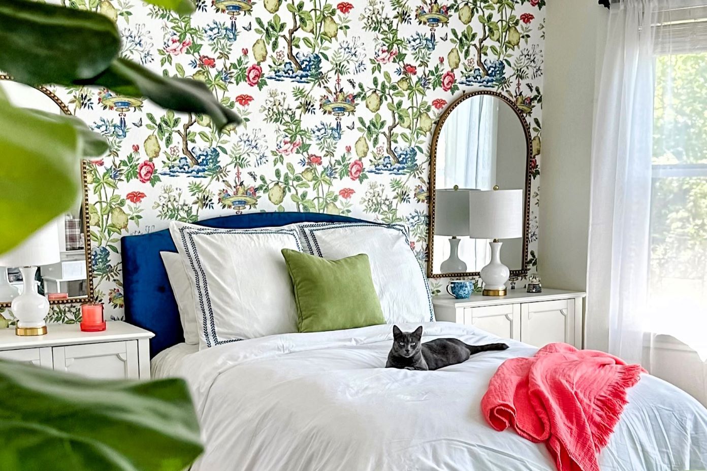 A bedroom with floral wallpaper, a blue velvet headboard, and a gray cat on the bed.
