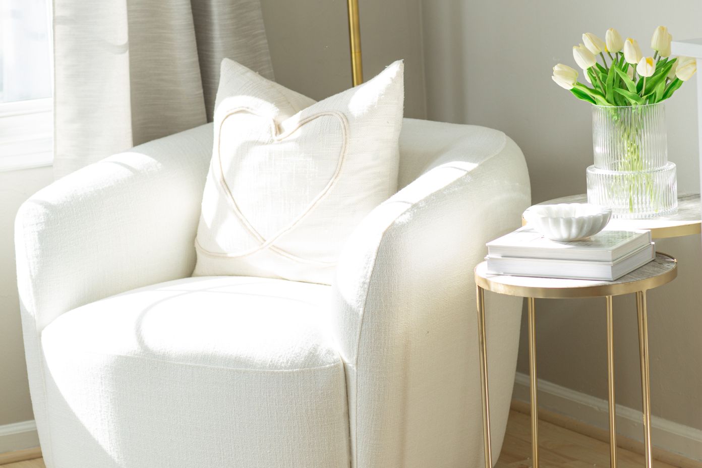 White armchair with a heart-shaped pillow, a side table with books, a bowl, and tulips.
