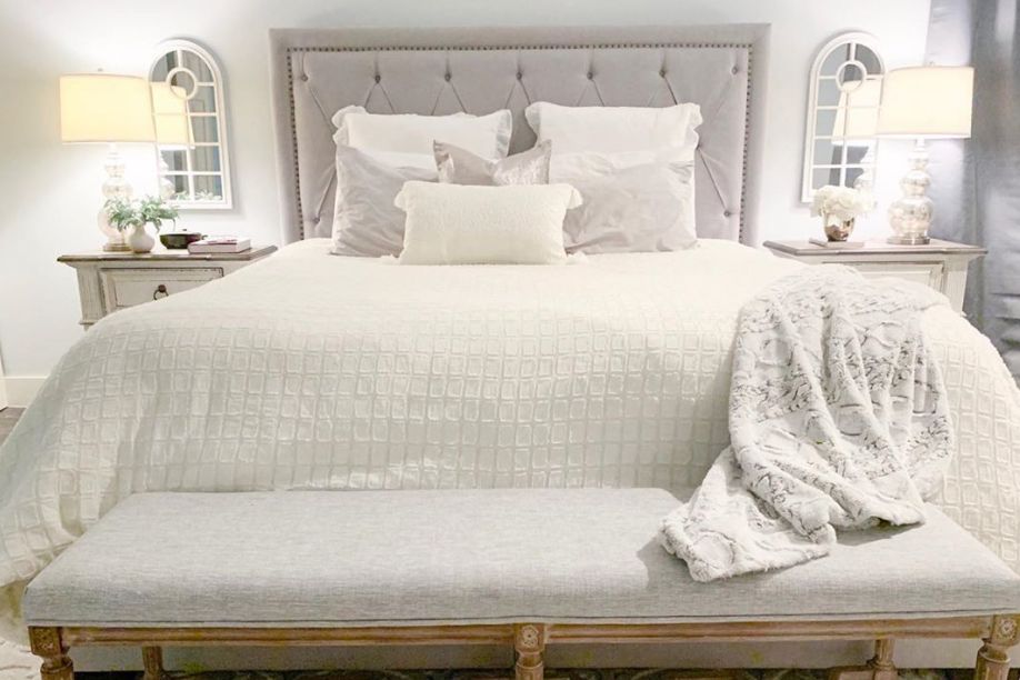 A bedroom with a bed, bench, and nightstands. The bed has a gray headboard and white bedding.
