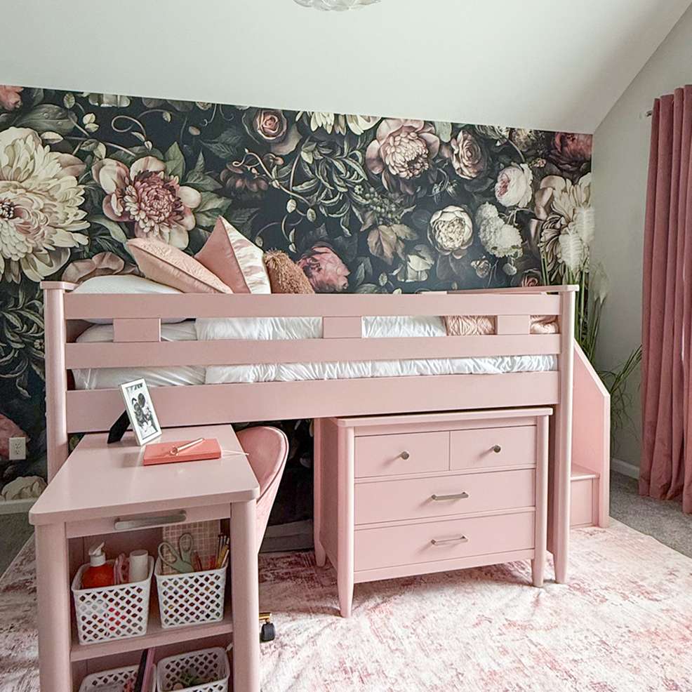 Pink loft bed with a desk and dresser, set against a floral wallpaper.
