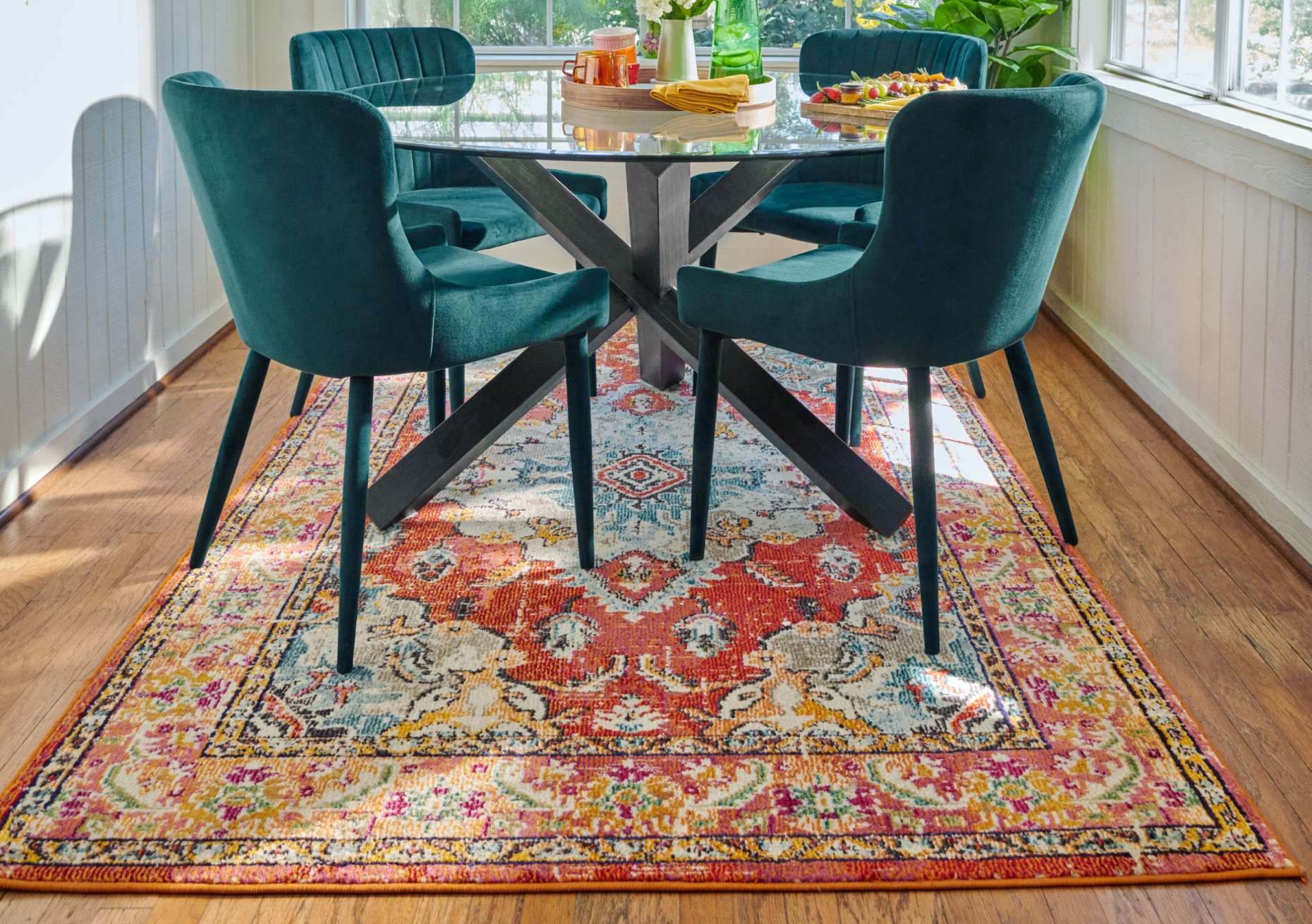 A dining room set with a round glass table, teal chairs, and a colorful rug.

