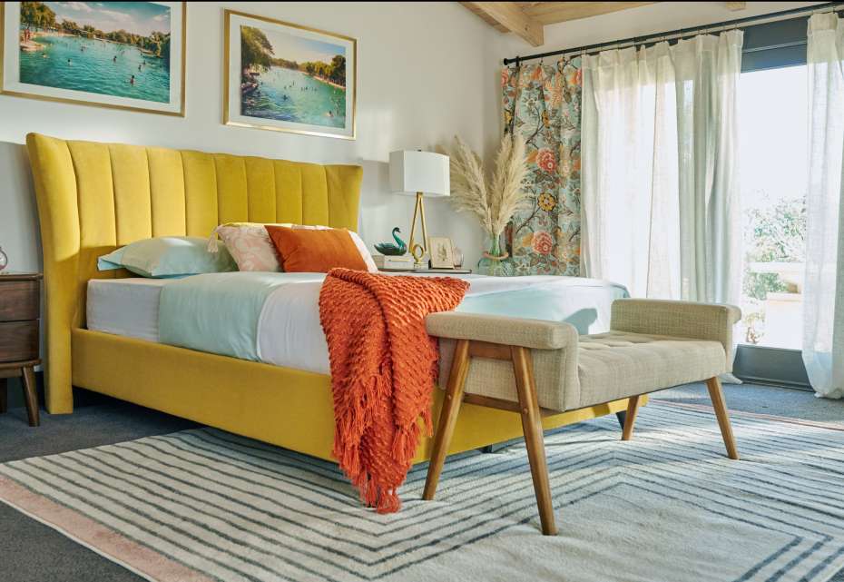 Yellow bed with orange throw blanket, bench, and patterned rug.
