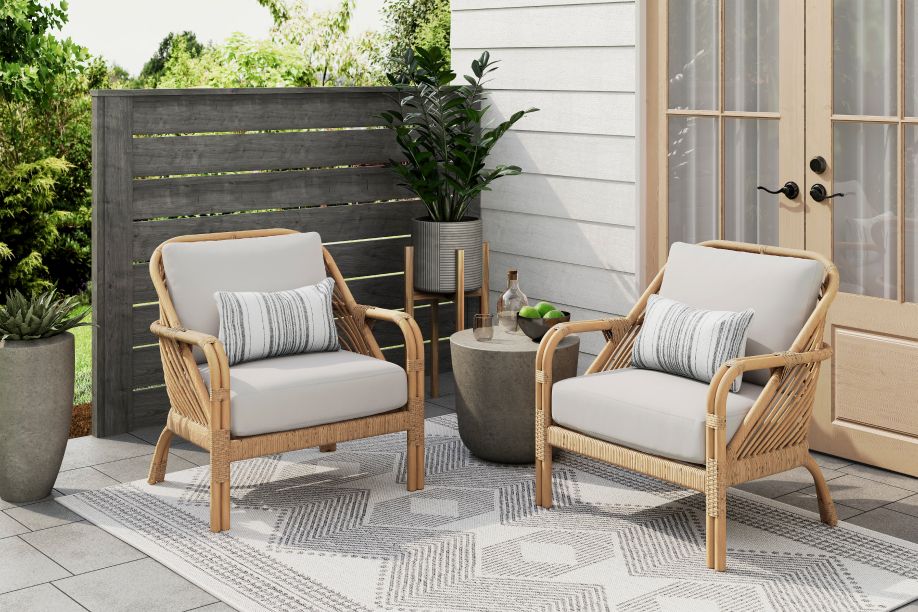Two wicker armchairs with light gray cushions and striped pillows on a patio with a rug and plants.
