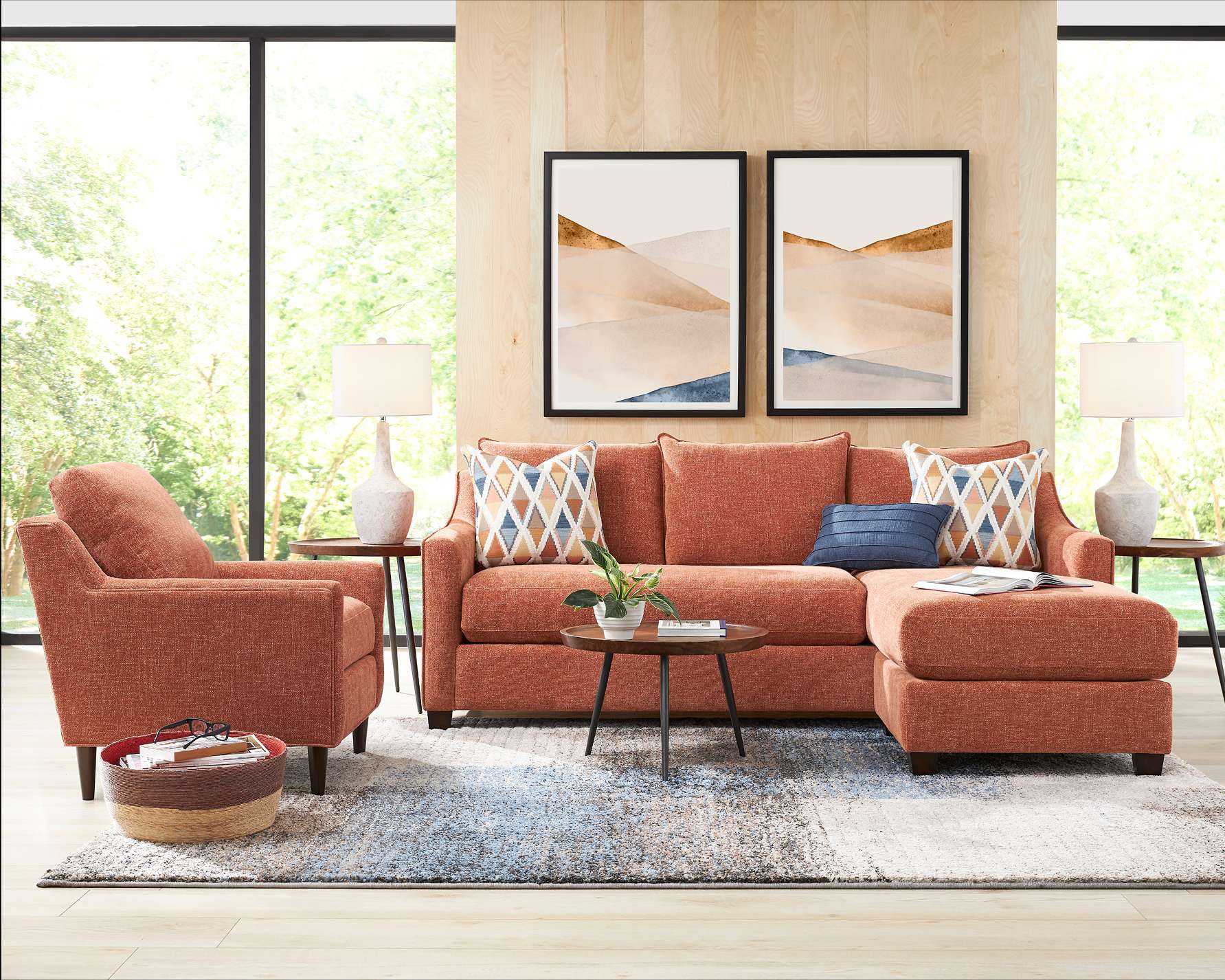 A living room with an orange sectional sofa, armchair, and rug. Two framed art pieces hang on the wall.
