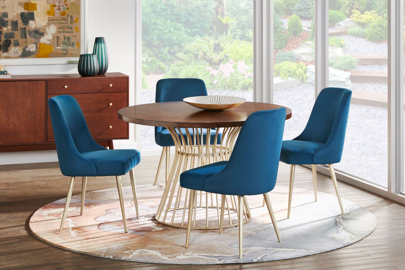 A dining room set with a round wooden table, blue velvet chairs, and a patterned rug.
