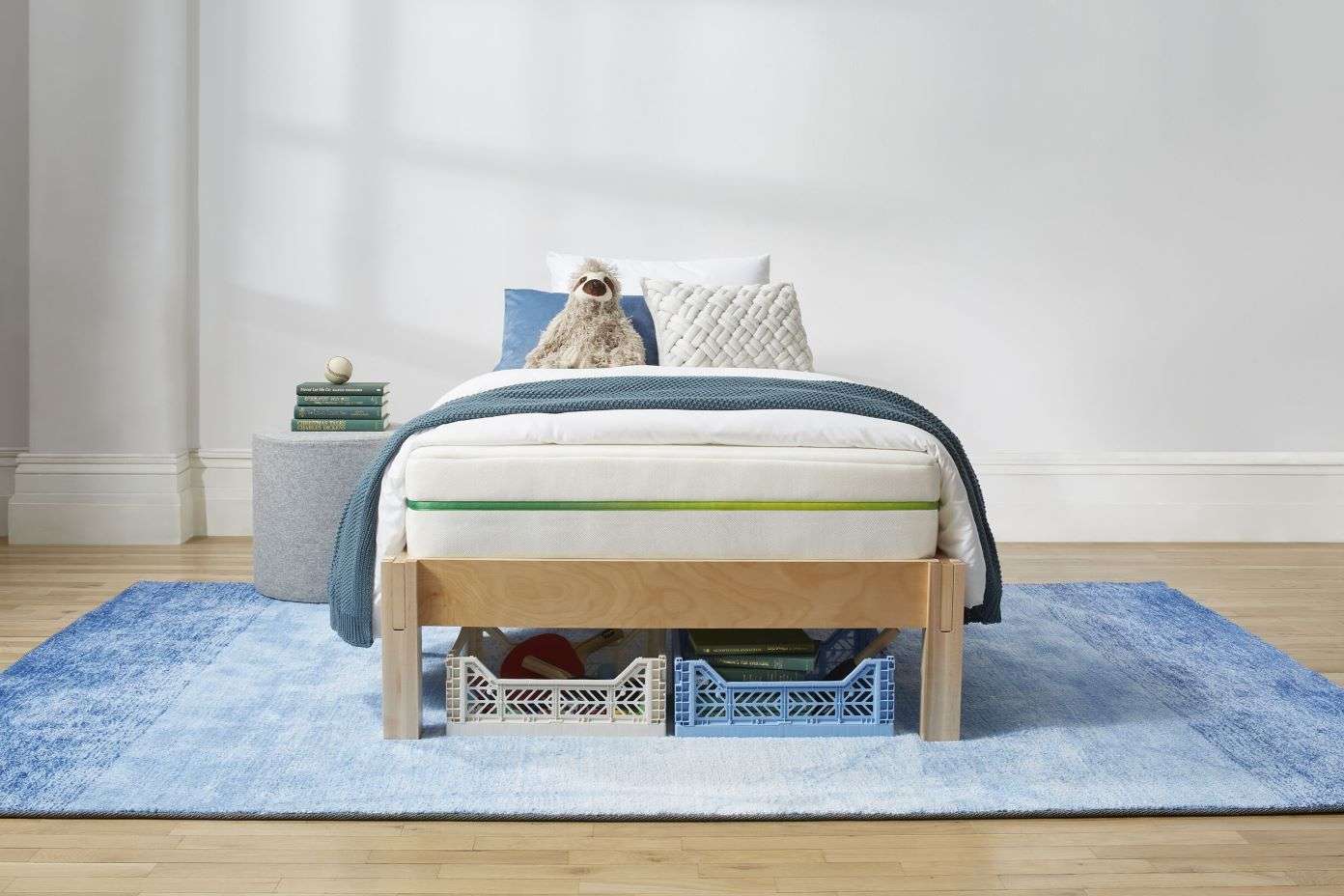 A bed with a mattress, pillows, and a blue blanket. Two storage crates are under the bed. A blue rug is on the floor.