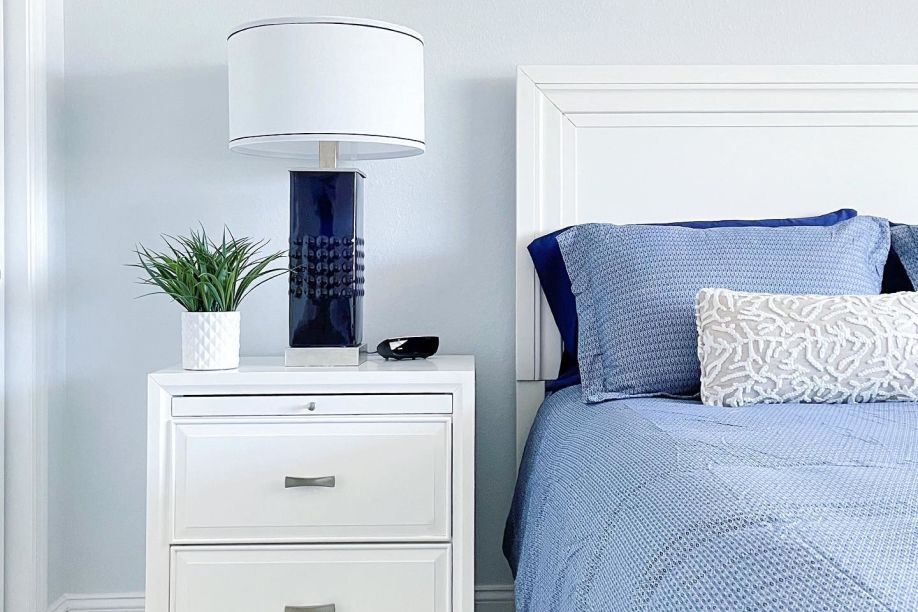 A white nightstand with a blue lamp, a plant, and a bed with blue bedding.
