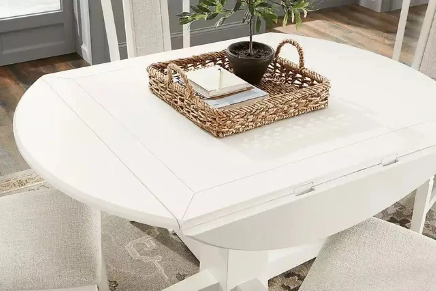 White drop-leaf dining table with a woven tray, plant, and notebooks.
