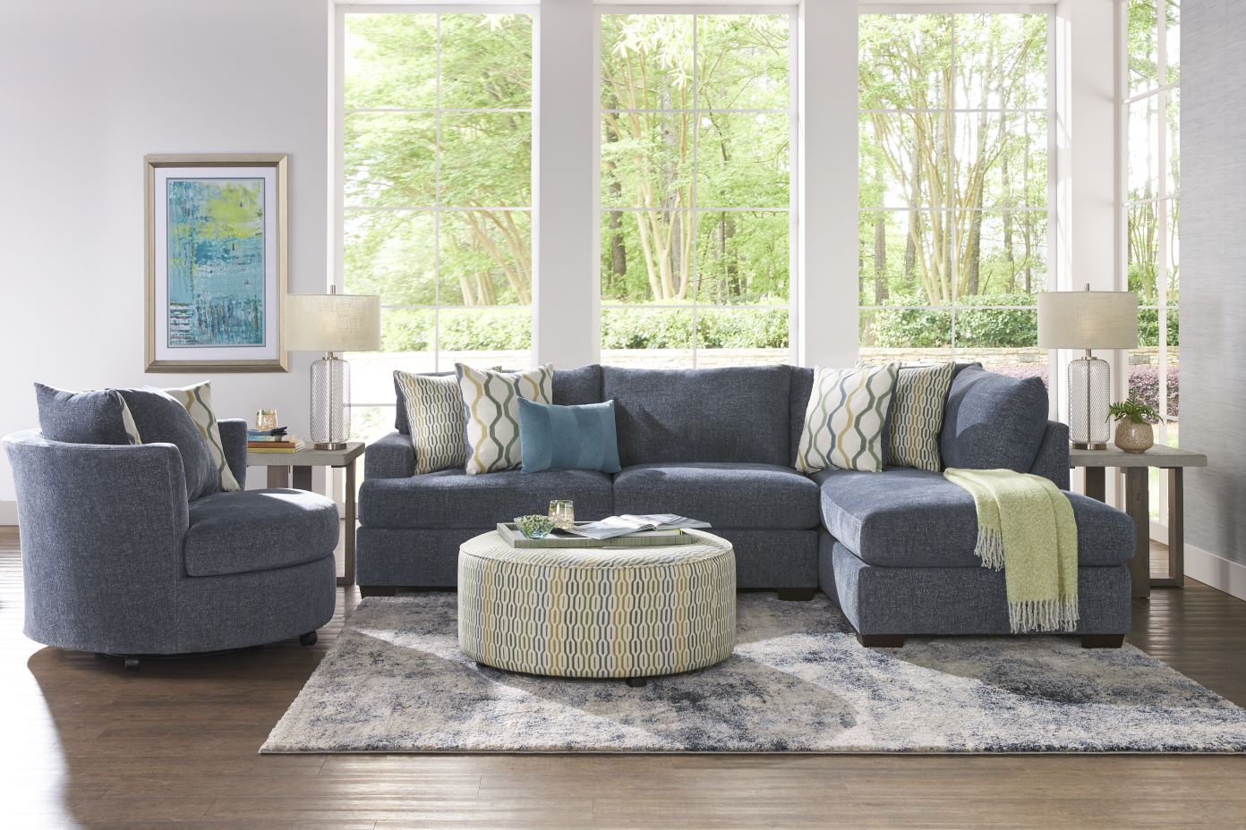 A living room with a blue sectional sofa, a round patterned ottoman, and a blue swivel chair.

