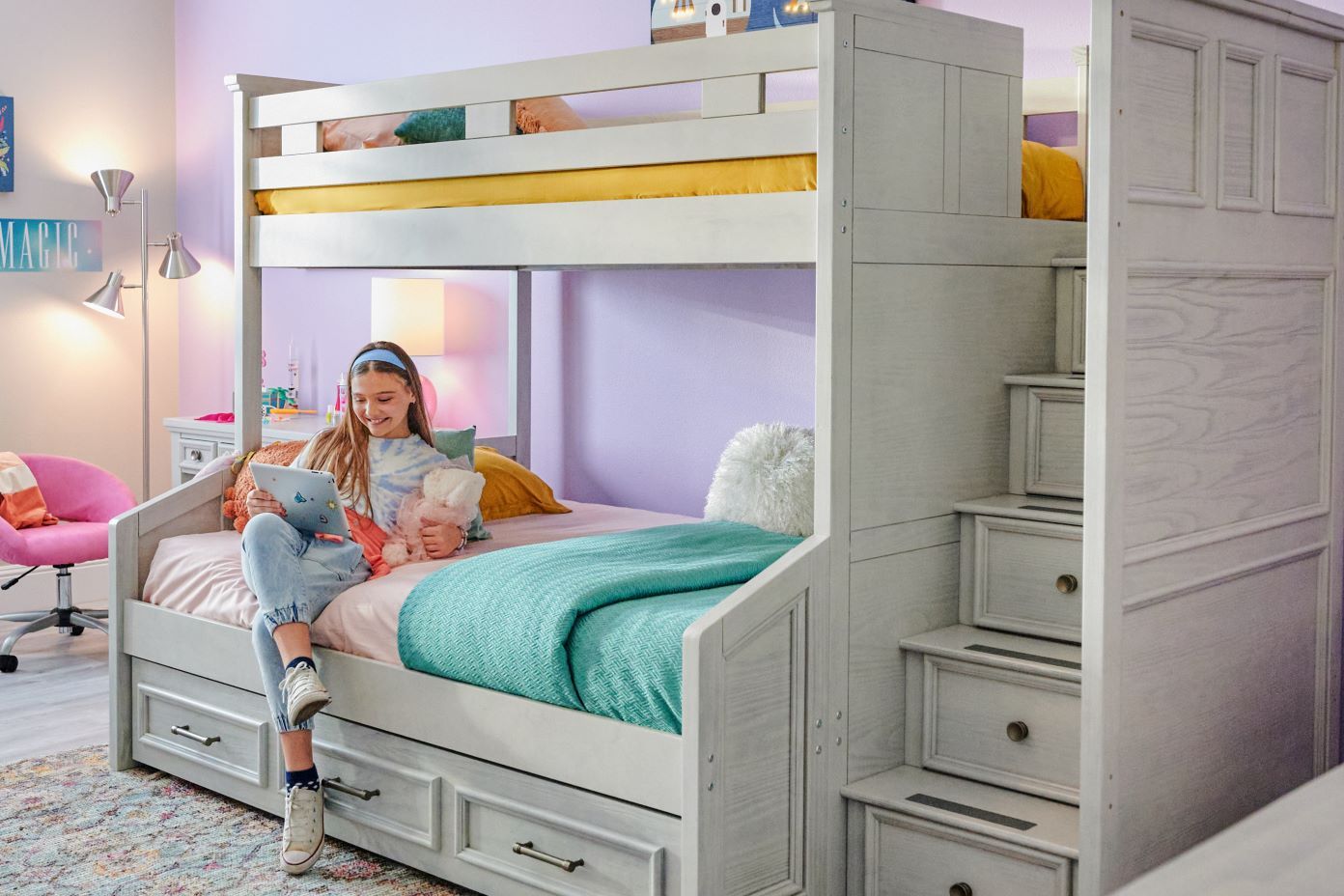 A girl sits on a bunk bed, holding a tablet and a stuffed animal. The bed has a staircase with drawers.
