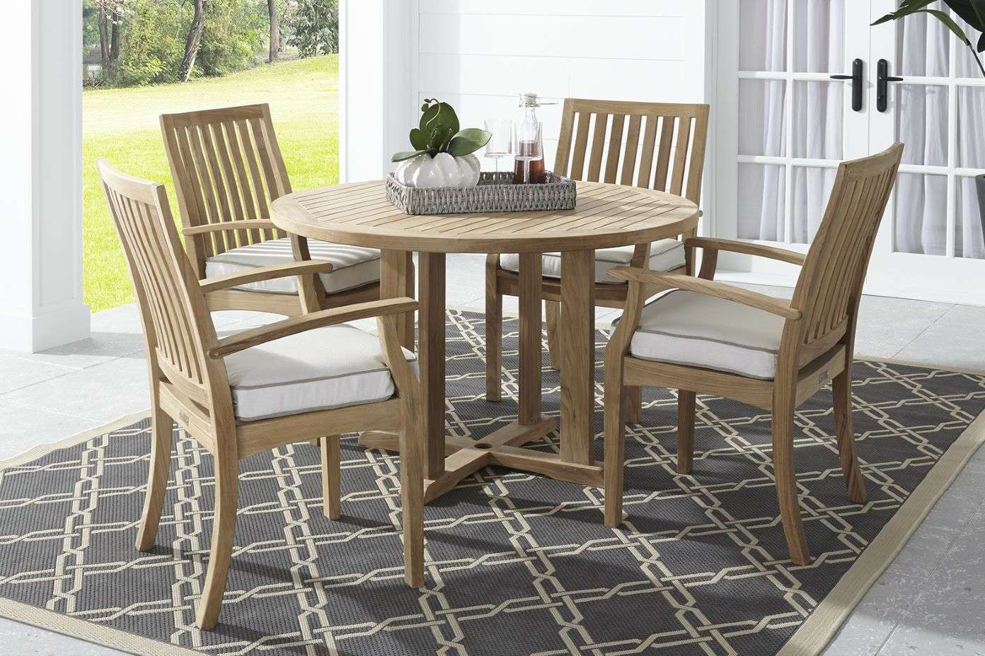Outdoor dining set with a round wooden table and four chairs with white cushions on a patterned rug.
