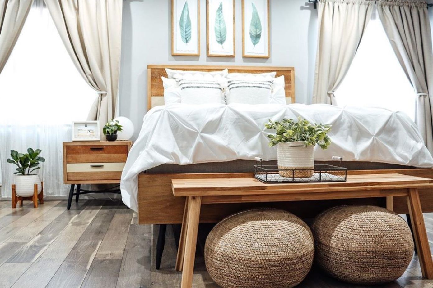 Bedroom with a wooden bed, bench, and two woven poufs. White bedding and curtains.
