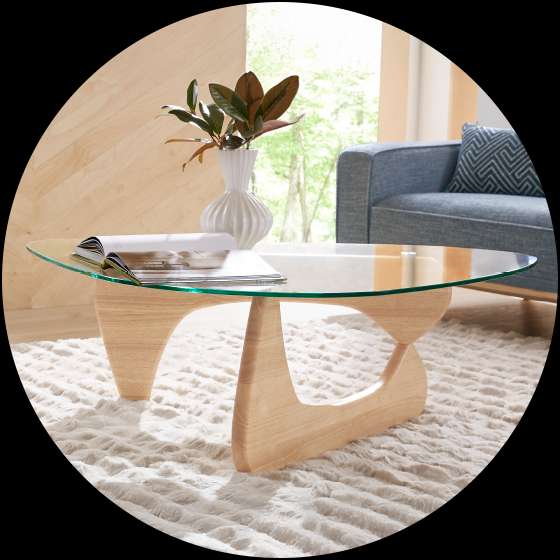 A modern coffee table with a glass top and wooden base, on a fluffy white rug.
