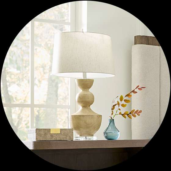 A wooden lamp with a beige lampshade sits on a dark brown nightstand. A vase with leaves is next to it.
