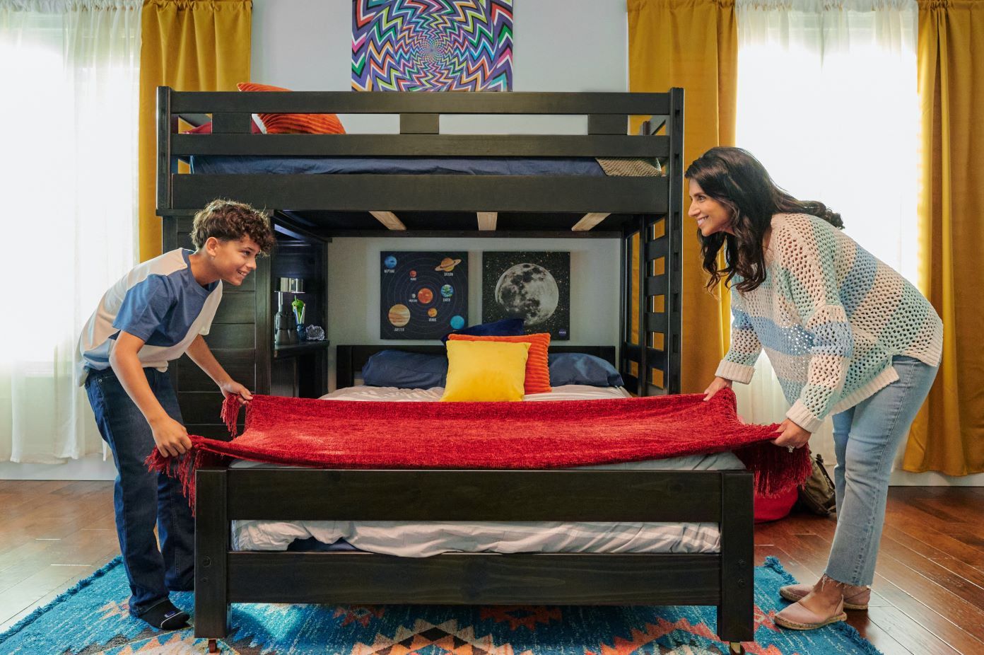 A woman and a boy are making a bed with a red blanket. The bed is a bunk bed.
