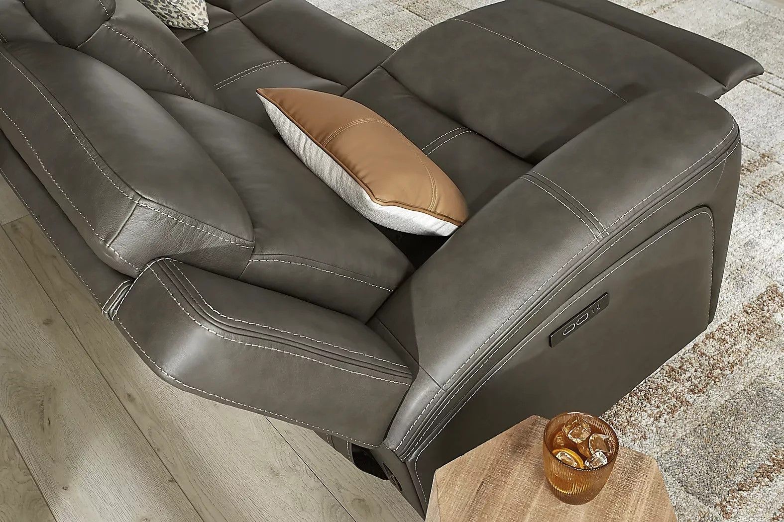Gray leather reclining sofa with white stitching and a brown pillow.
