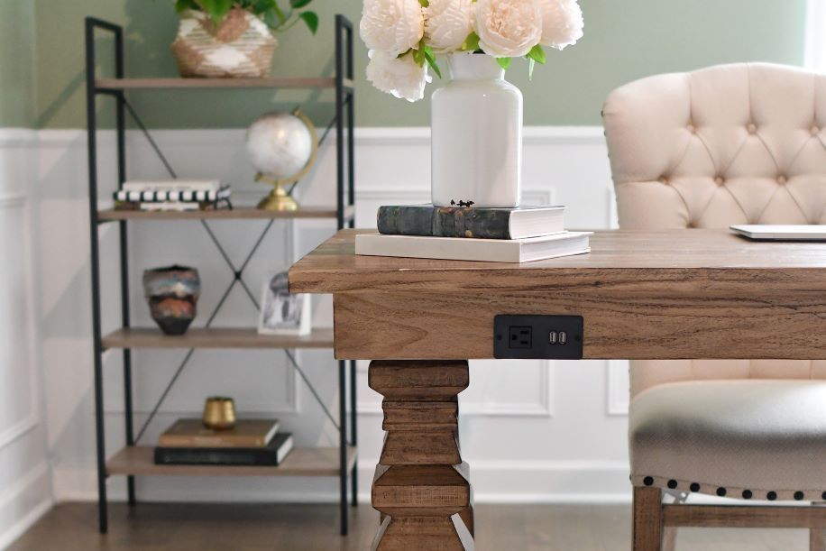 Wooden desk with a white vase of flowers, books, and a chair. A bookshelf is in the background.

