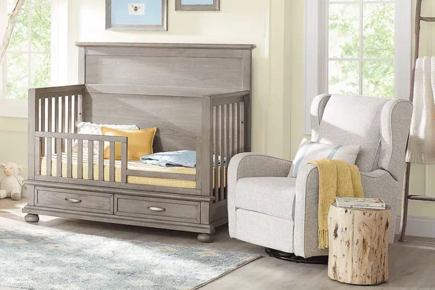 A gray crib with two drawers, a gray rocking chair, and a tree stump side table.
