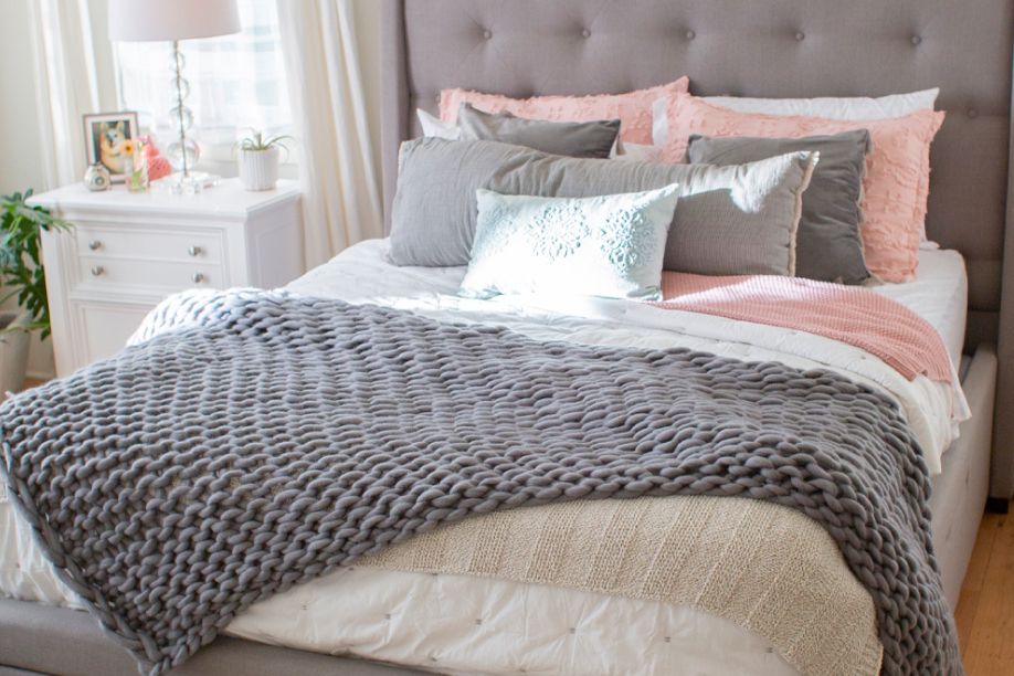 A bed with a gray knitted blanket, pillows, and a white nightstand.
