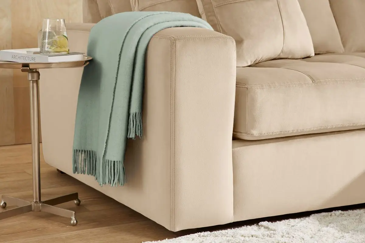 A beige upholstered sofa with a light green throw blanket draped over the arm, next to a small side table with a book.