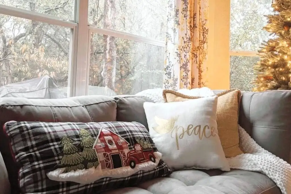 A cozy living room with a gray couch and Christmas pillows. A window looks out onto a wooded area.
