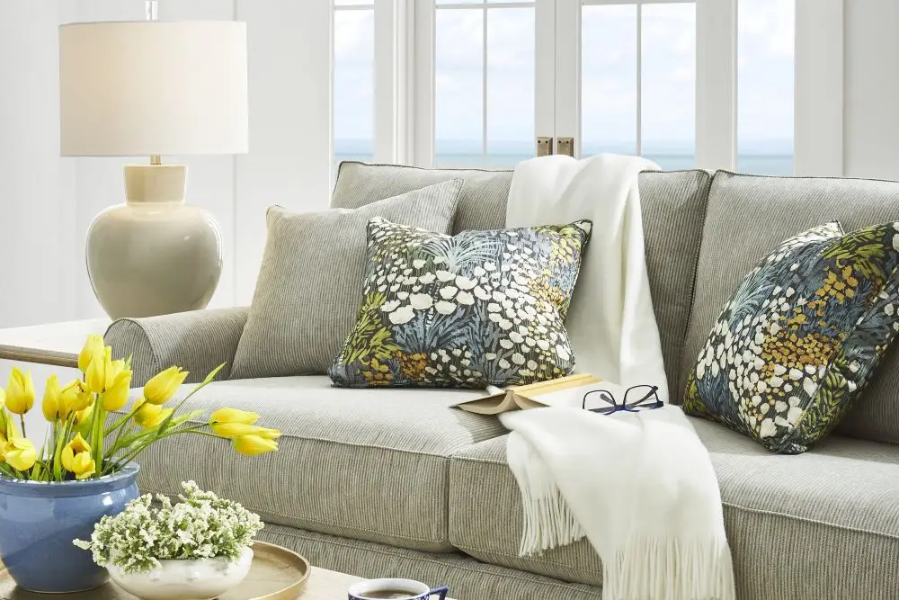 A gray sofa with floral accent pillows, a white throw blanket, a book, glasses, and a vase of yellow tulips on a table.