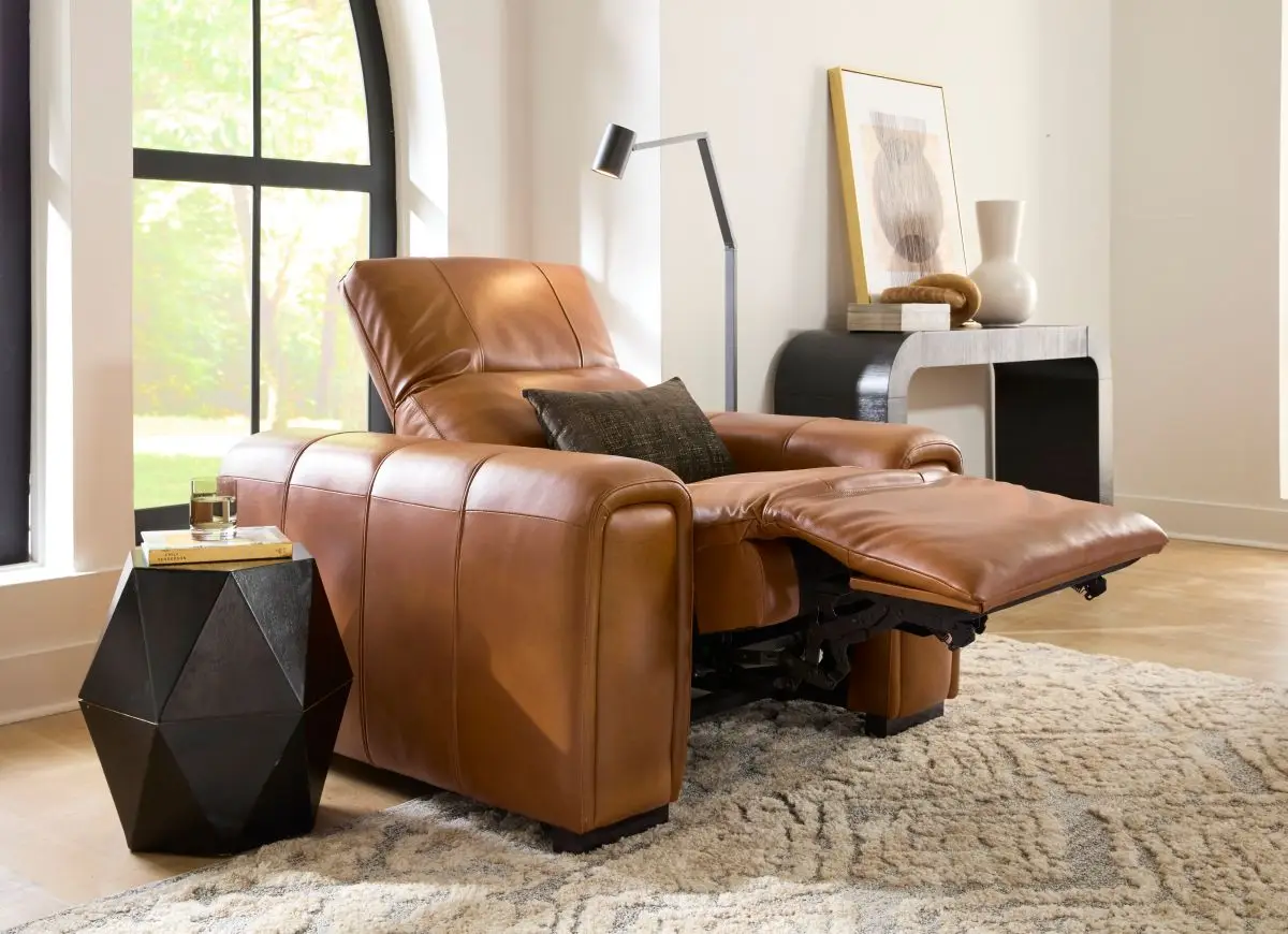 A tan leather recliner chair with a dark accent pillow, positioned next to a black geometric side table in a bright room.