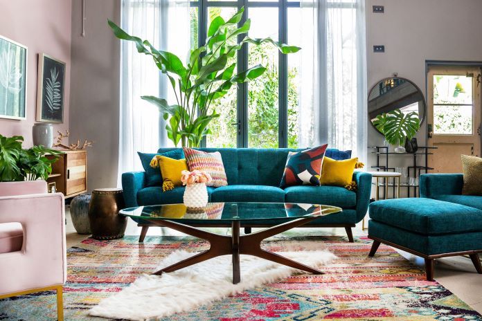 A teal sofa with colorful pillows, a glass coffee table, and a patterned rug.
