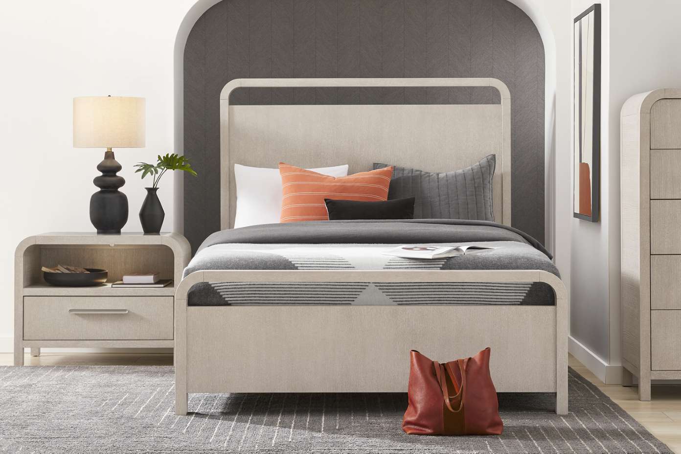 Bedroom with a bed, nightstand, and dresser. The bed has a gray blanket and orange pillow. A red tote bag sits on the floor.