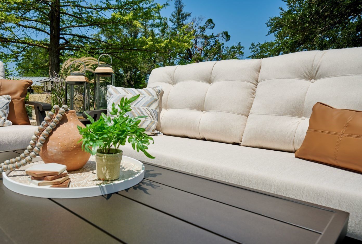 Outdoor sofa with cream cushions and brown leather pillows. A tray with a plant and decorative items sits on the table.
