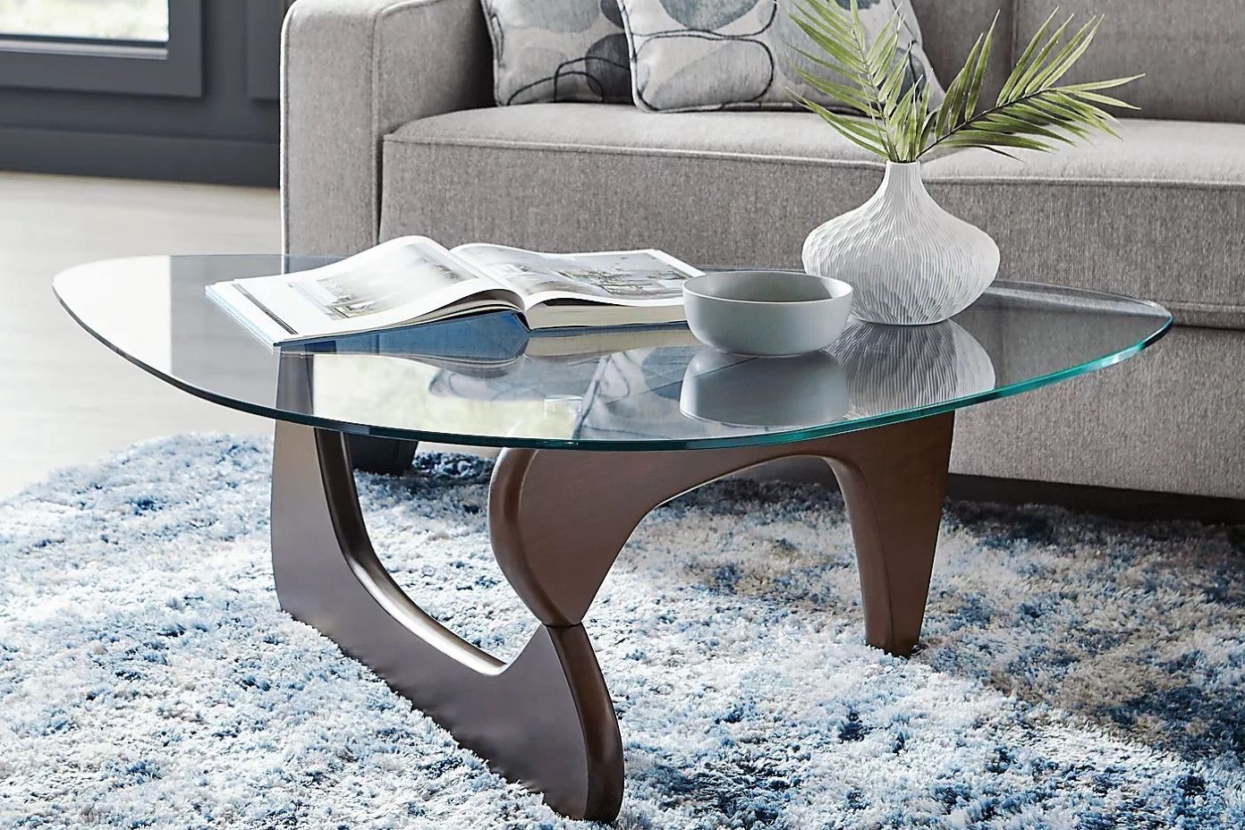 Glass coffee table with a wooden base, a book, a bowl, and a vase with greenery.

