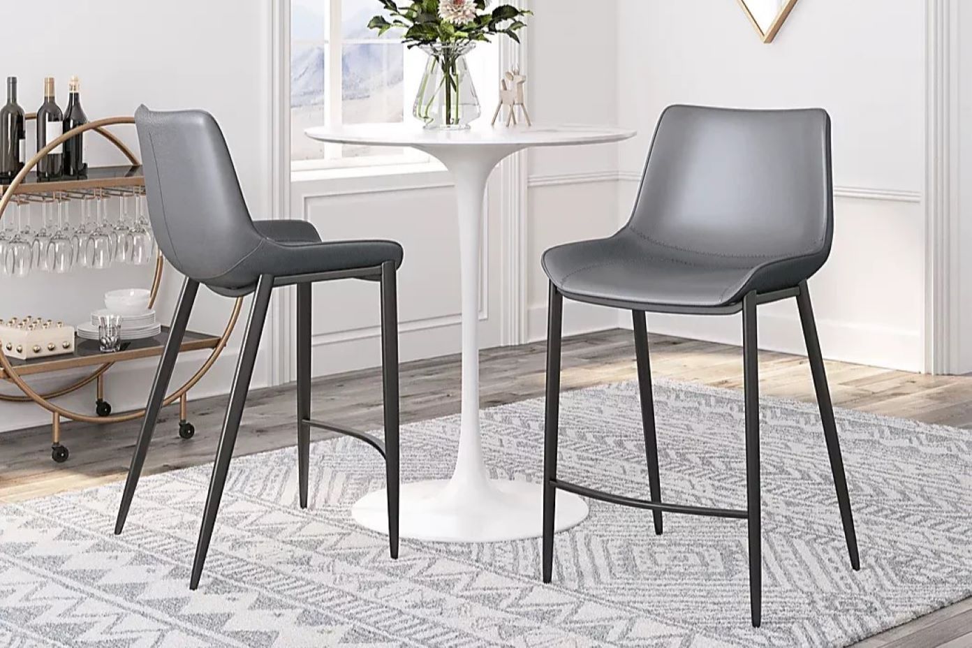 Two gray bar stools with black legs sit on a patterned rug near a white table.
