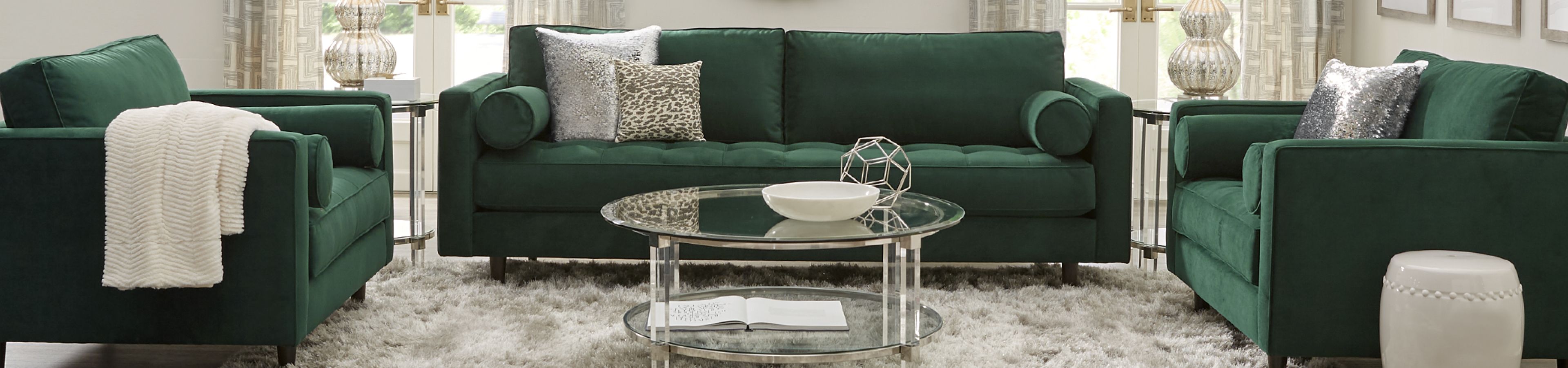 Green velvet sofa and armchairs with decorative pillows, a glass coffee table, and a white rug.
