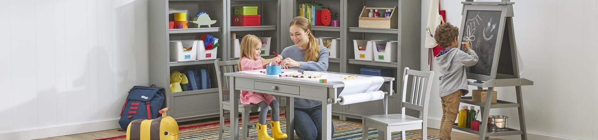 A woman and a young girl sit at a table, drawing. A boy draws on a chalkboard.
