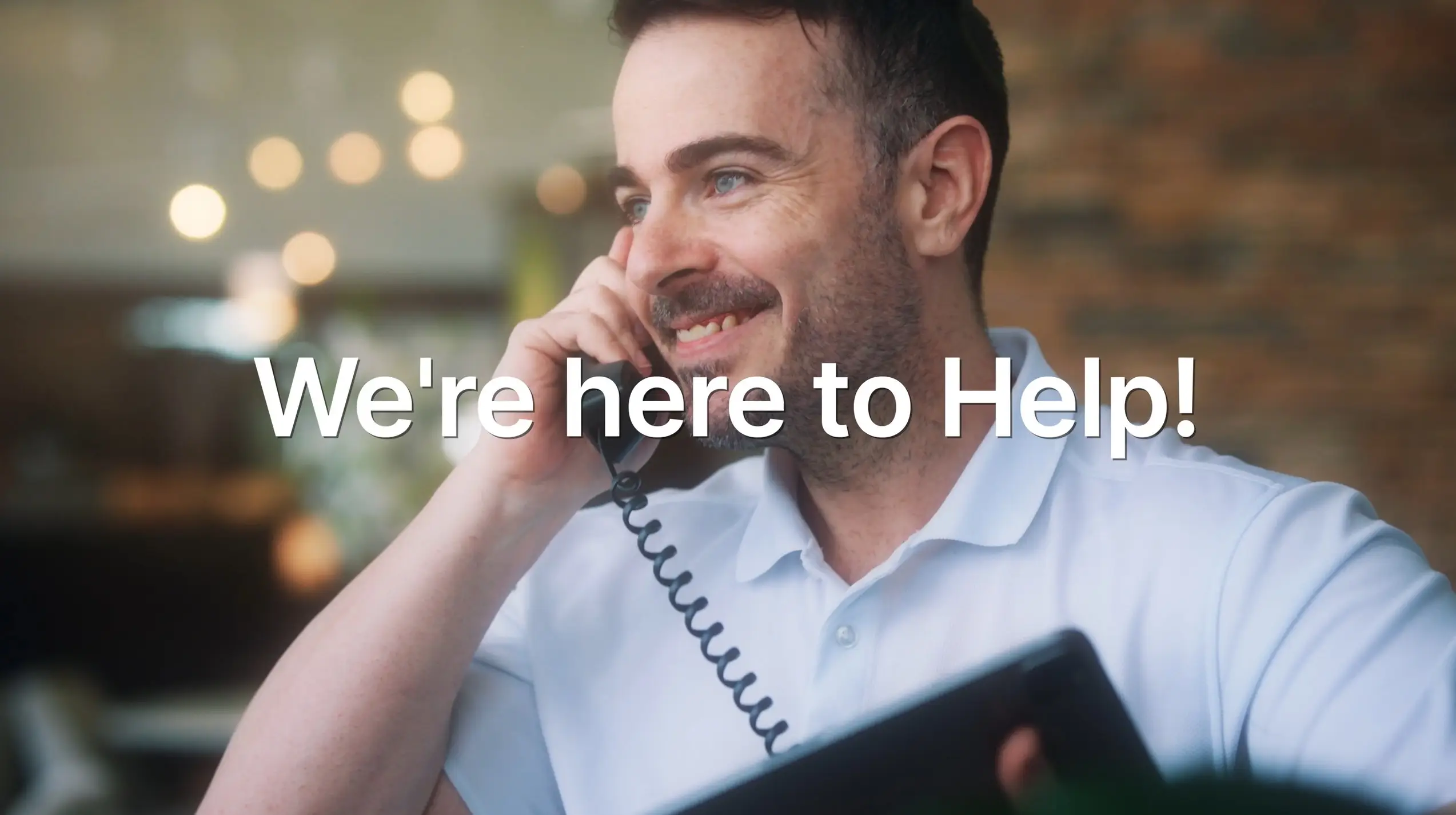 We're here to Help! A smiling man in a white polo shirt talks on a landline phone while holding a tablet.