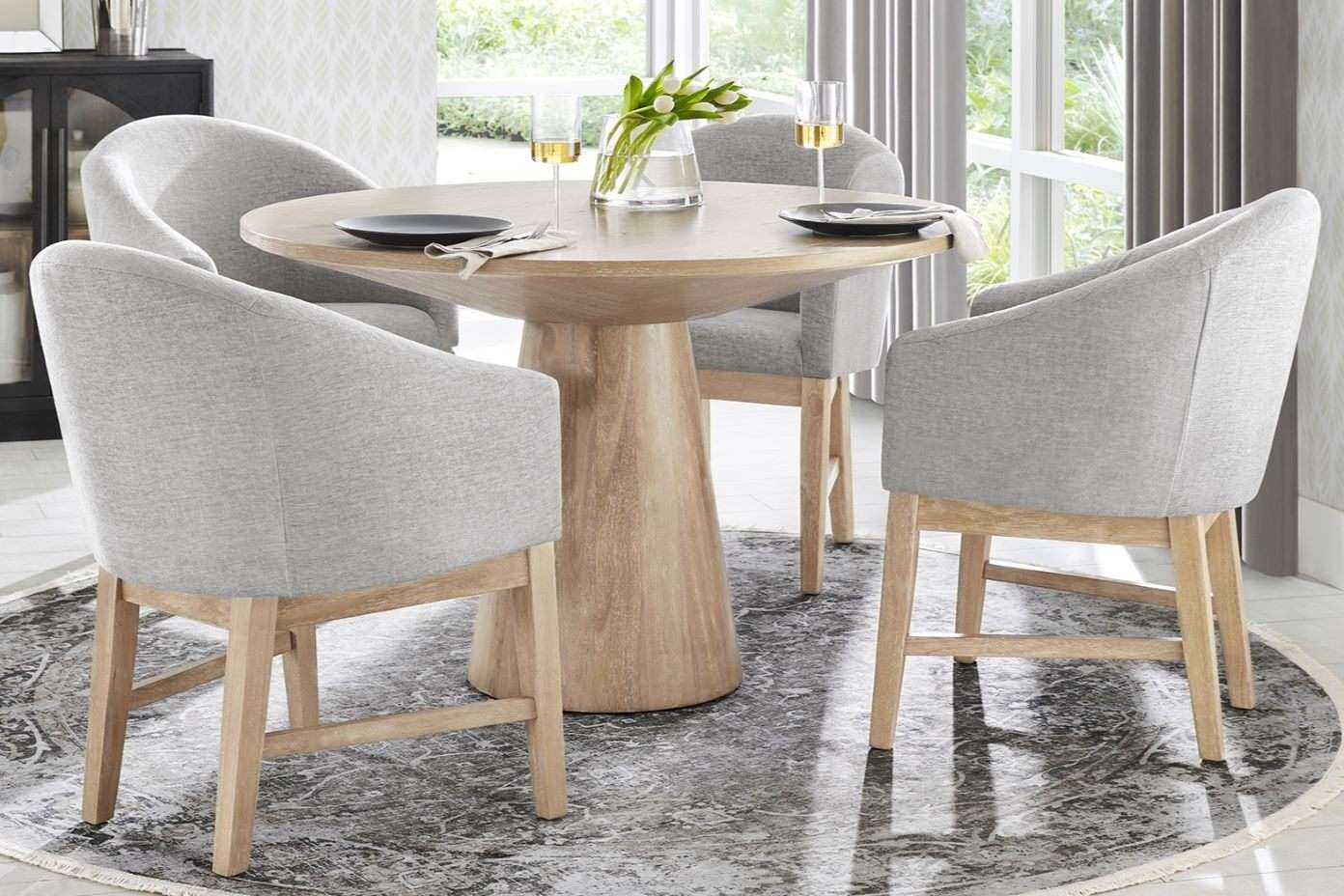 A round wooden dining table with four gray upholstered chairs on a patterned rug.
