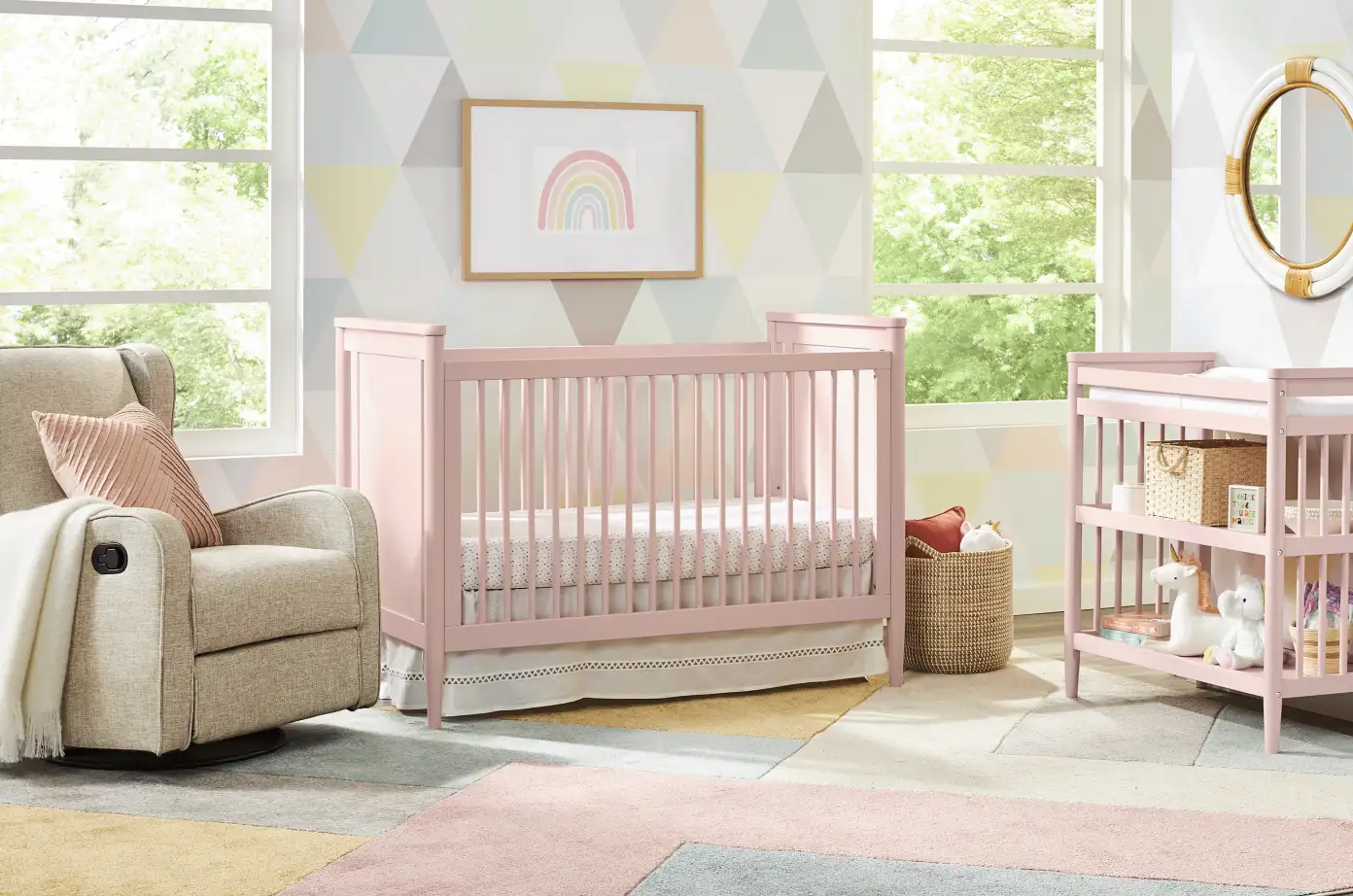 Pink crib and changing table in a nursery with a rainbow picture and a rocking chair.
