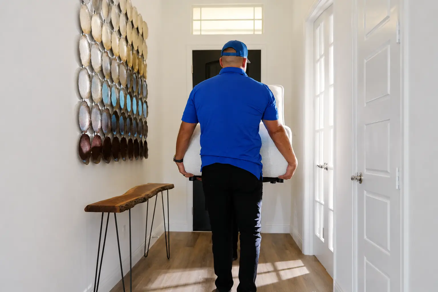 A mover in a blue uniform carries a white piece of furniture through a hallway with a decorative wall art piece.