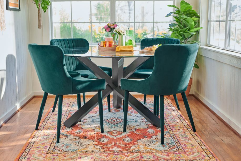 A dining room set with a glass table, teal chairs, and a patterned rug.
